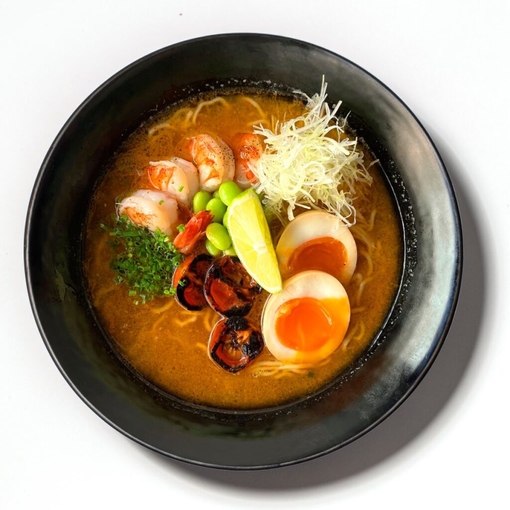 Ramen de camarão servido em taça preta com ovo marinado, edamame e caldo intenso
