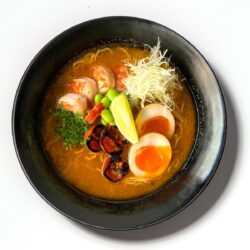 Ramen de camarão servido em taça preta com ovo marinado, edamame e caldo intenso